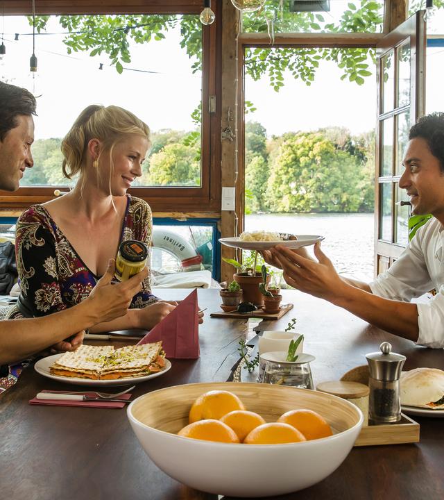 "Matze, Kebab und Sauerkraut": Noah (Franz Dinda), Charlottes (Christine Eixenberger) und Hakim (Omar El-Saeidi) sitzen gemeinsam am Tisch. Hakim hält Charlotte einen Teller hin, im Hintergrund glitzert der See