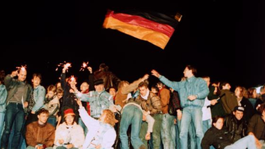 Menschen feiern auf der Berliner Mauer und halten eine Deutschland-Fahne 