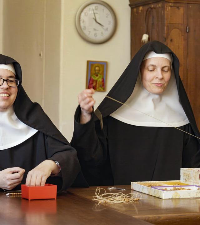 Das Bild zeigt zwei klosterbekleidete Frauen, die in einem Raum sitzen, der ein warmes, einladendes Ambiente ausstrahlt. Beide tragen traditionelle Habitbekleidung, bestehend aus schwarzen Gewändern und weißen Hauben. Die Frau auf der linken Seite hat eine Brille und lächelt, während sie an einem kleinen roten Kästchen sitzt. Ihre Hände sind beschäftigt, wahrscheinlich mit einer Art Bastelarbeit oder Hobby. Die andere Frau, die sich rechts befindet, hält einen Faden in der Hand und sieht konzentriert aus.