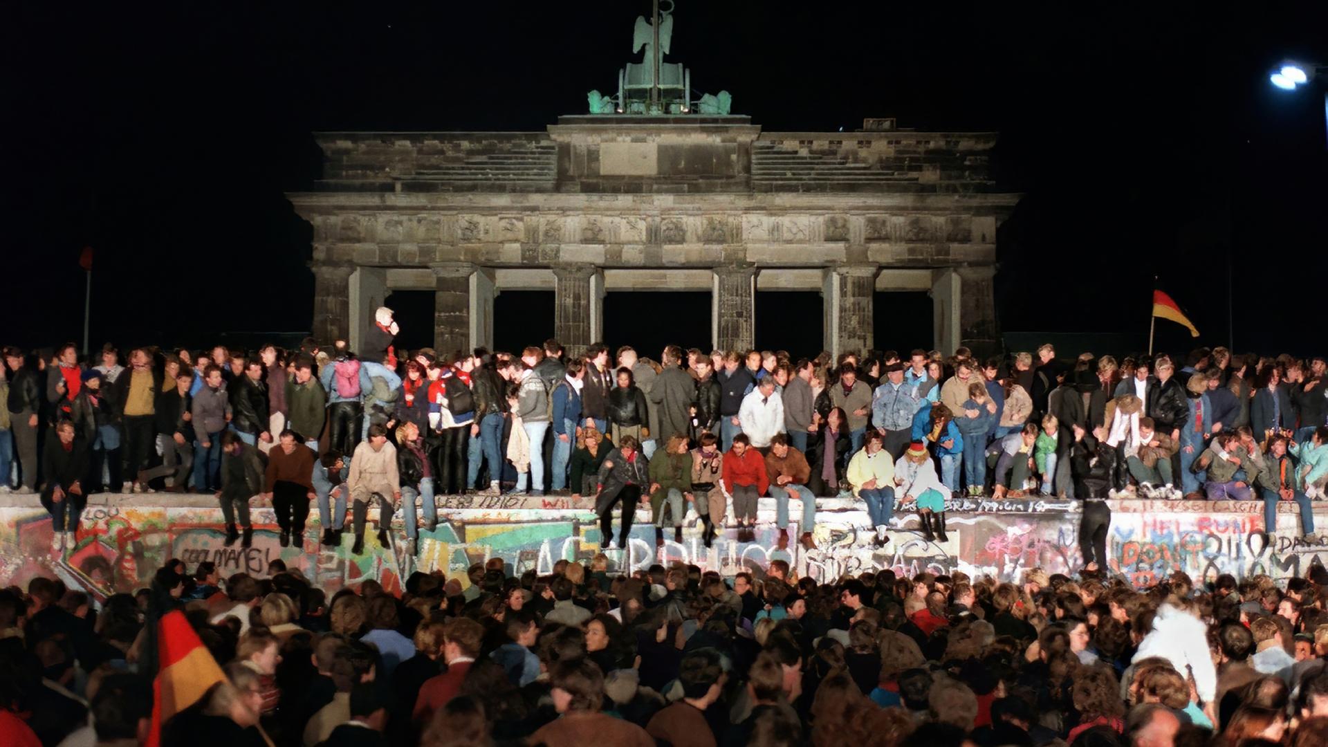 Auf der bunt bemalten Betonmauer vor dem Brandenburger Tor in Berlin sitzen und stehen Menschen. Davor hat sich eine Menschenmenge versammelt.