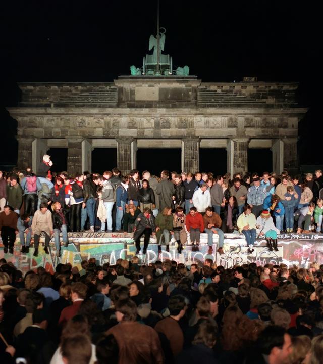 Auf der bunt bemalten Betonmauer vor dem Brandenburger Tor in Berlin sitzen und stehen Menschen. Davor hat sich eine Menschenmenge versammelt.