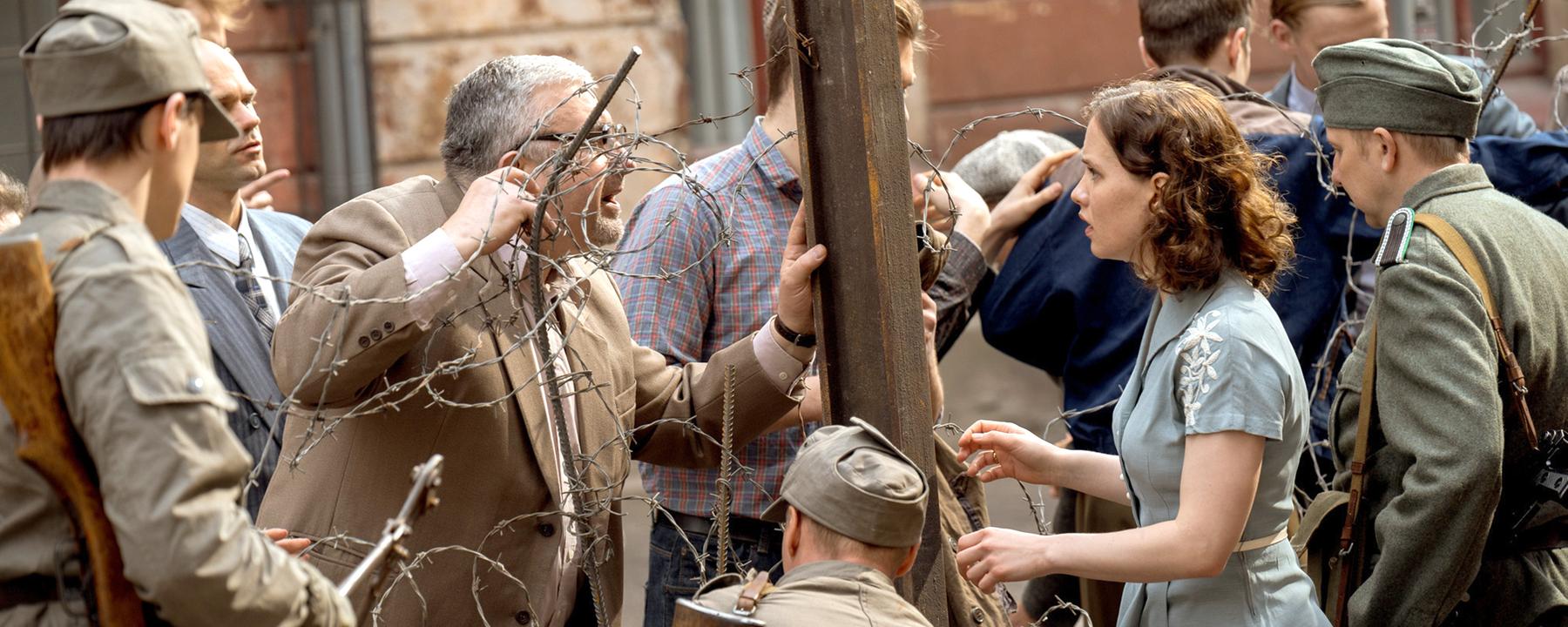 Menschen, getrennt durch Stacheldraht, darunter eine junge Frau, Ingrid Taegner (Sandija Dovgāne), zwischen Grenzsoldaten, die einen älteren Mann auf der anderen Seite des provisorischen Zauns anschaut.