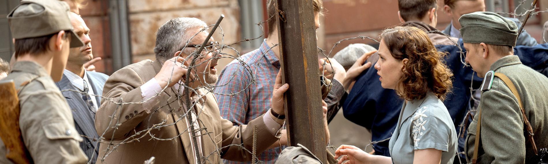 Menschen, getrennt durch Stacheldraht, darunter eine junge Frau, Ingrid Taegner (Sandija Dovgāne), zwischen Grenzsoldaten, die einen älteren Mann auf der anderen Seite des provisorischen Zauns anschaut.
