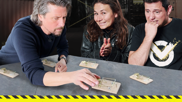Tommy Scheel, Maja und Cem sitzen an einem Tisch mit mehreren Mausefallen, auf denen Geldstücke platziert sind. Maja und Cem grinsen schelmisch, Tommy versucht vorsichtig Geld von der Falle zu nehmen.