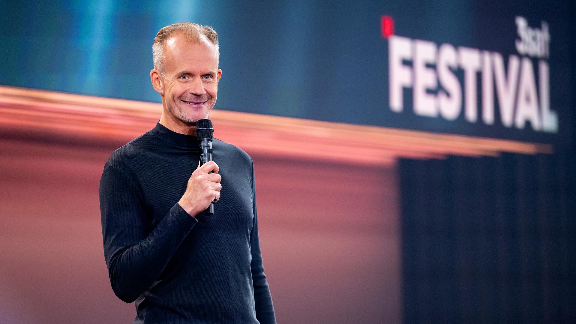 Max Uthoff steht auf der linken Seite des Bildes. Er hält ein Mikrofon in seiner rechten Hand. Die Bühne hinter ihm ist unten rot und oben blau beleuchtet. Max Uthoff ist schwarz gekleidet.