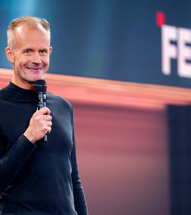 Max Uthoff steht auf der linken Seite des Bildes. Er hält ein Mikrofon in seiner rechten Hand. Die Bühne hinter ihm ist unten rot und oben blau beleuchtet. Max Uthoff ist schwarz gekleidet.