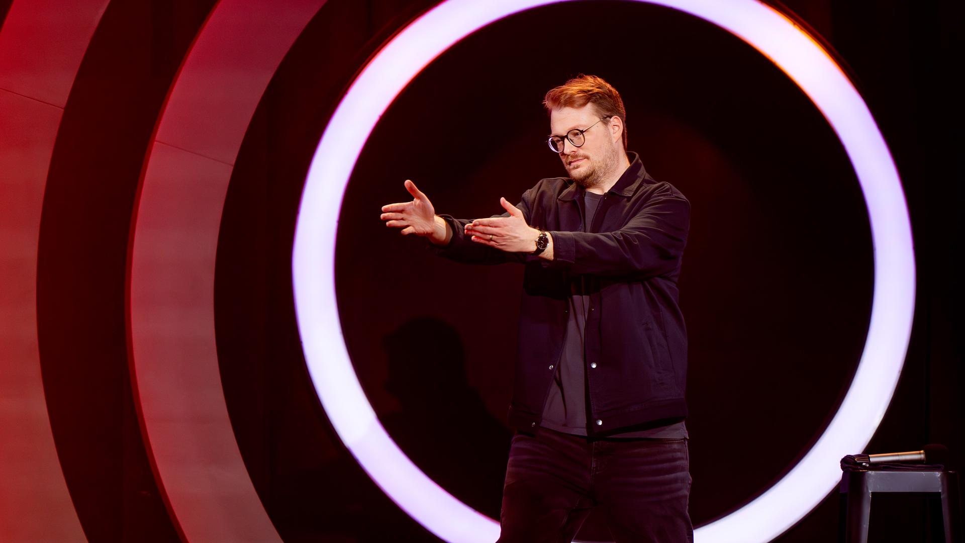 Maxi Gstettenbauer steht auf der rechten Bildseite. Er streckt beide Arme in Richtung des linken Bildrandes aus. Er ist dunkel gekleidet. Auf der rechten Seite steht ein schwarzer Barhocker, auf dem ein Mikrofon liegt. Maxi Gstettenbauer trägt eine Brille.