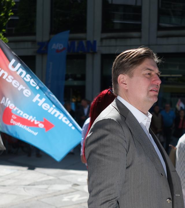 Ein Mann in Anzug läuft auf der Straße, im Hintergrund trägt ein Mann eine Flagge, auf der Unser Land, unsere Heimat und Alternative für Deutschland steht.
