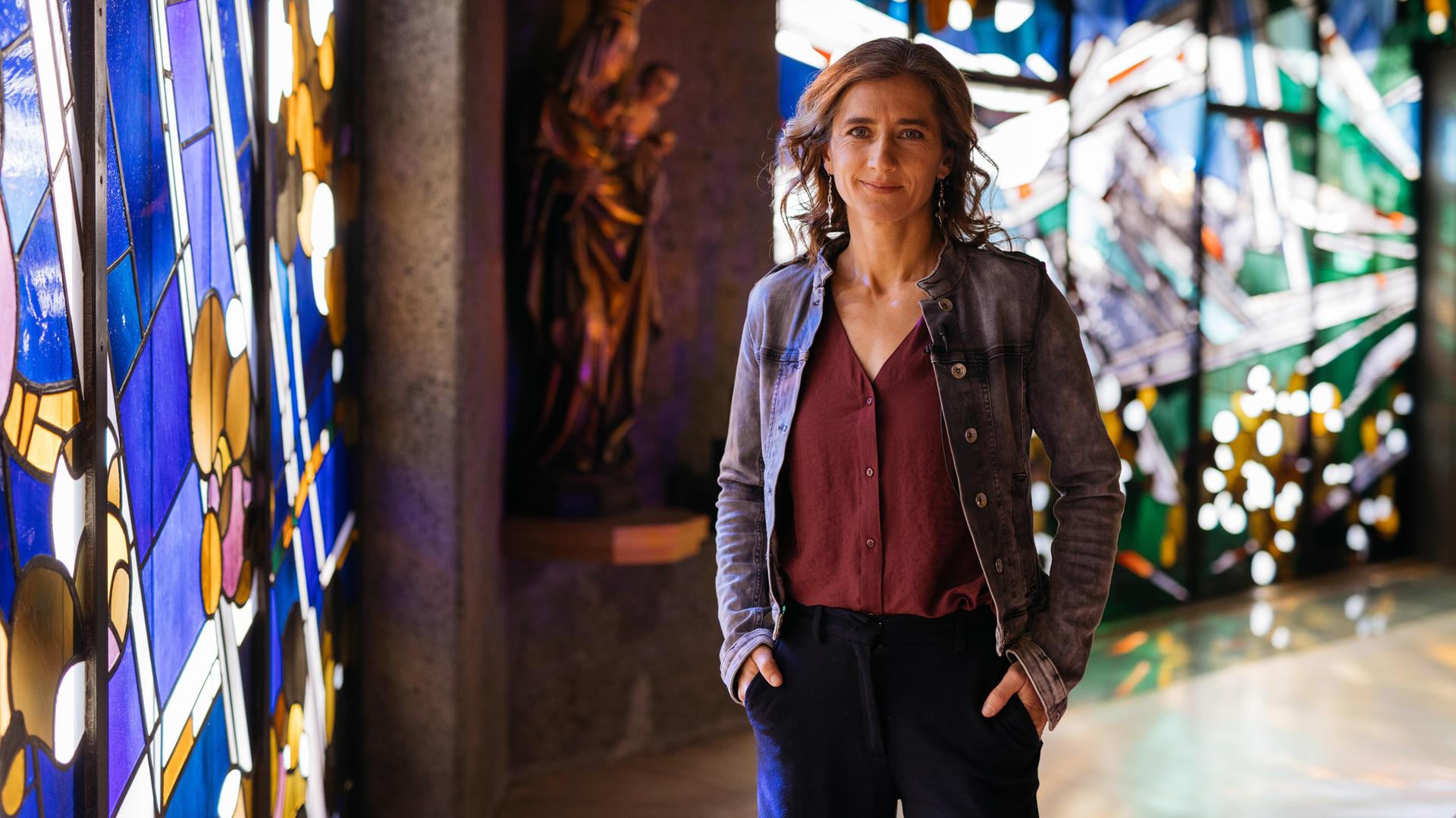 Elena steht in einer Kirche, die Hände lässig in die Hosentaschen gesteckt - sie lächelt in die Kamera. Im Hintergrund sieht man links eine Marienfigur, die das Jesuskind im Arm hält sowie bunte Kirchenfenster. 