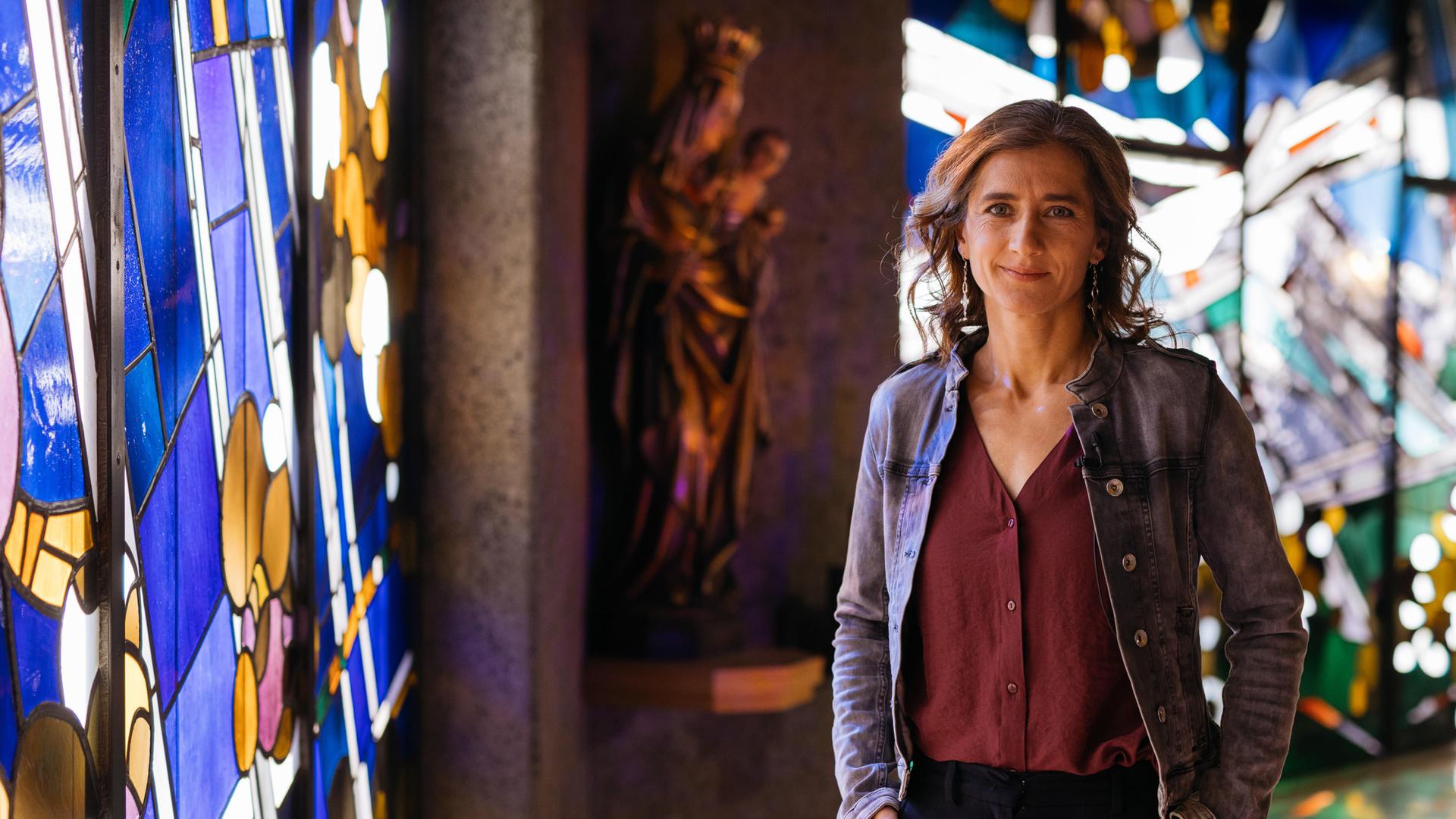 Elena steht in einer Kirche, die Hände lässig in die Hosentaschen gesteckt - sie lächelt in die Kamera. Im Hintergrund sieht man links eine Marienfigur, die das Jesuskind im Arm hält sowie bunte Kirchenfenster. 