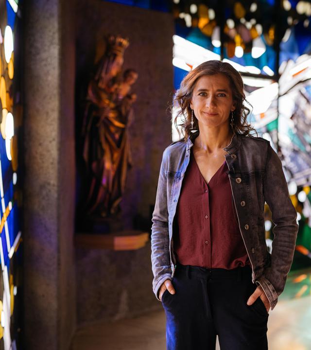 Elena steht in einer Kirche, die Hände lässig in die Hosentaschen gesteckt - sie lächelt in die Kamera. Im Hintergrund sieht man links eine Marienfigur, die das Jesuskind im Arm hält sowie bunte Kirchenfenster. 