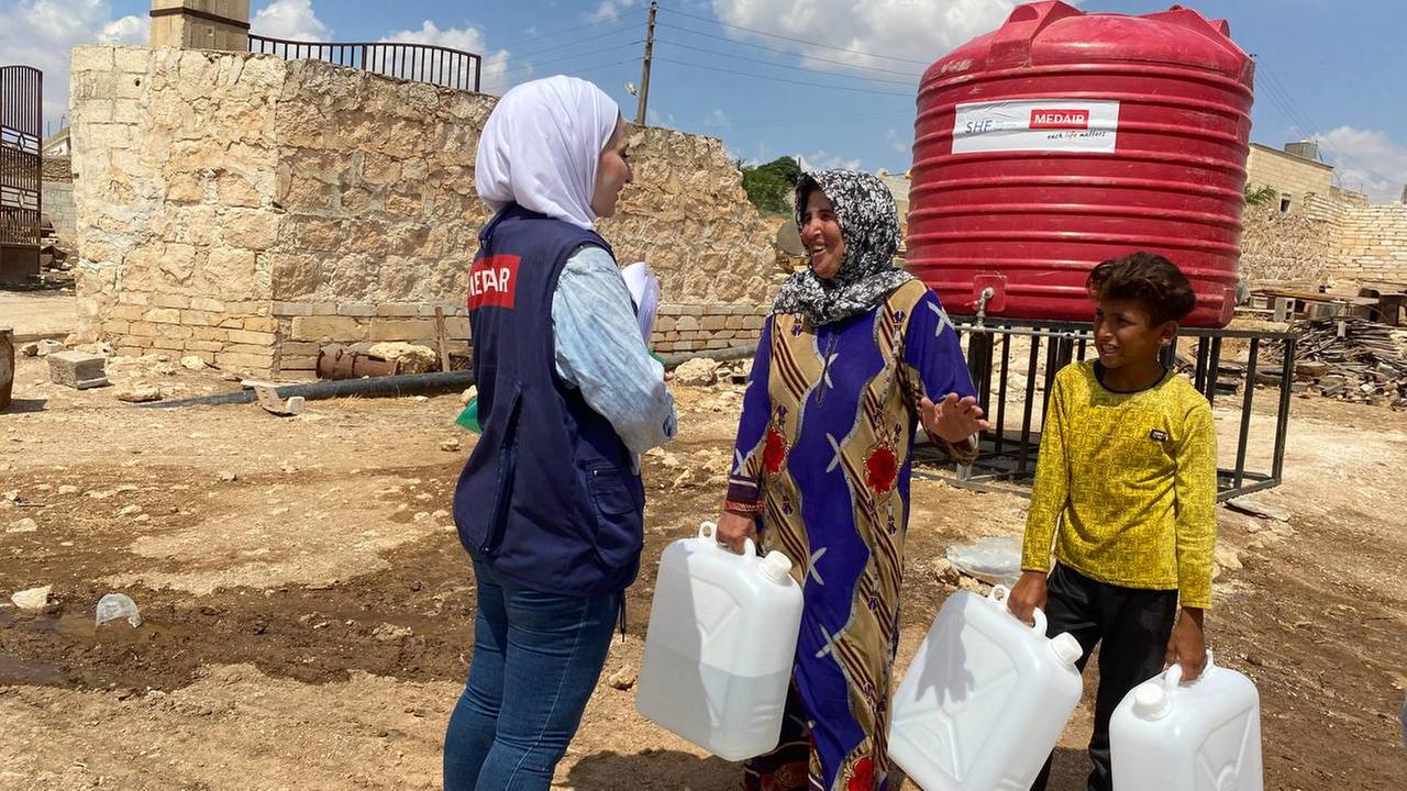 Zwei Kinder in Syrien mit Trinkwasserkanistern und eine Frau der Hilfsorganisation Medair
