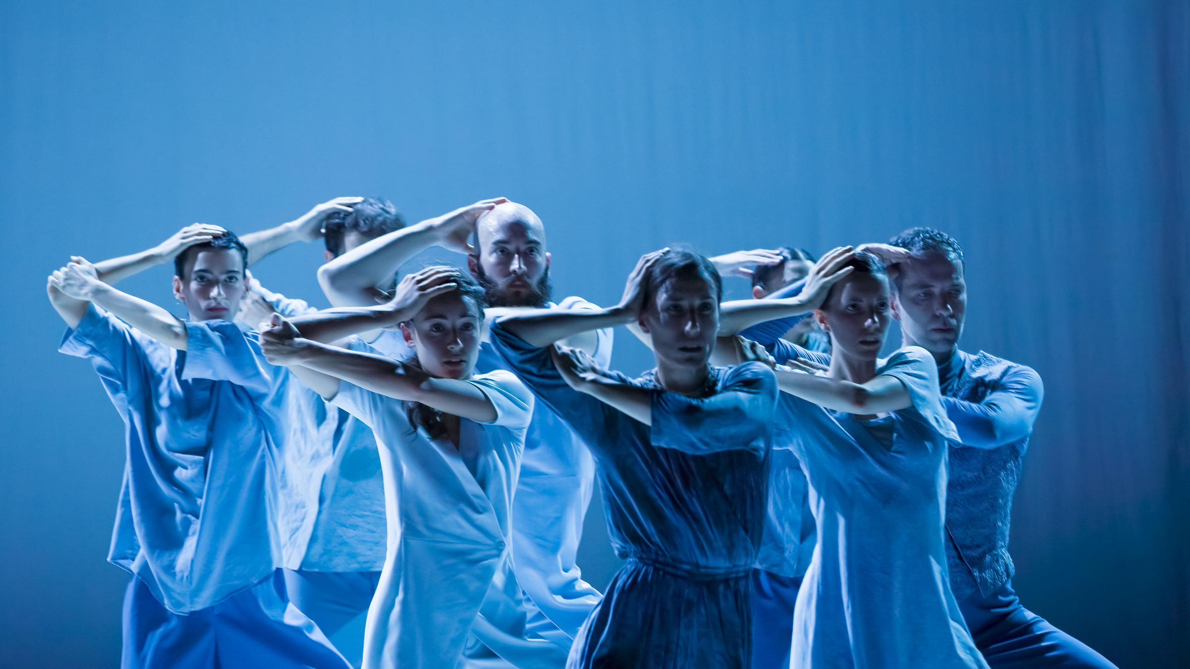 Eine Gruppe von fünf Tänzerinnen und drei Tänzern in blauen Kostümen, sie blicken gerade nach vorne, den rechten Arm angewinkelt und die Hand auf den eigenen Kopf gelegt, mit dem linken Arm den rechten Ellbogen haltend. 