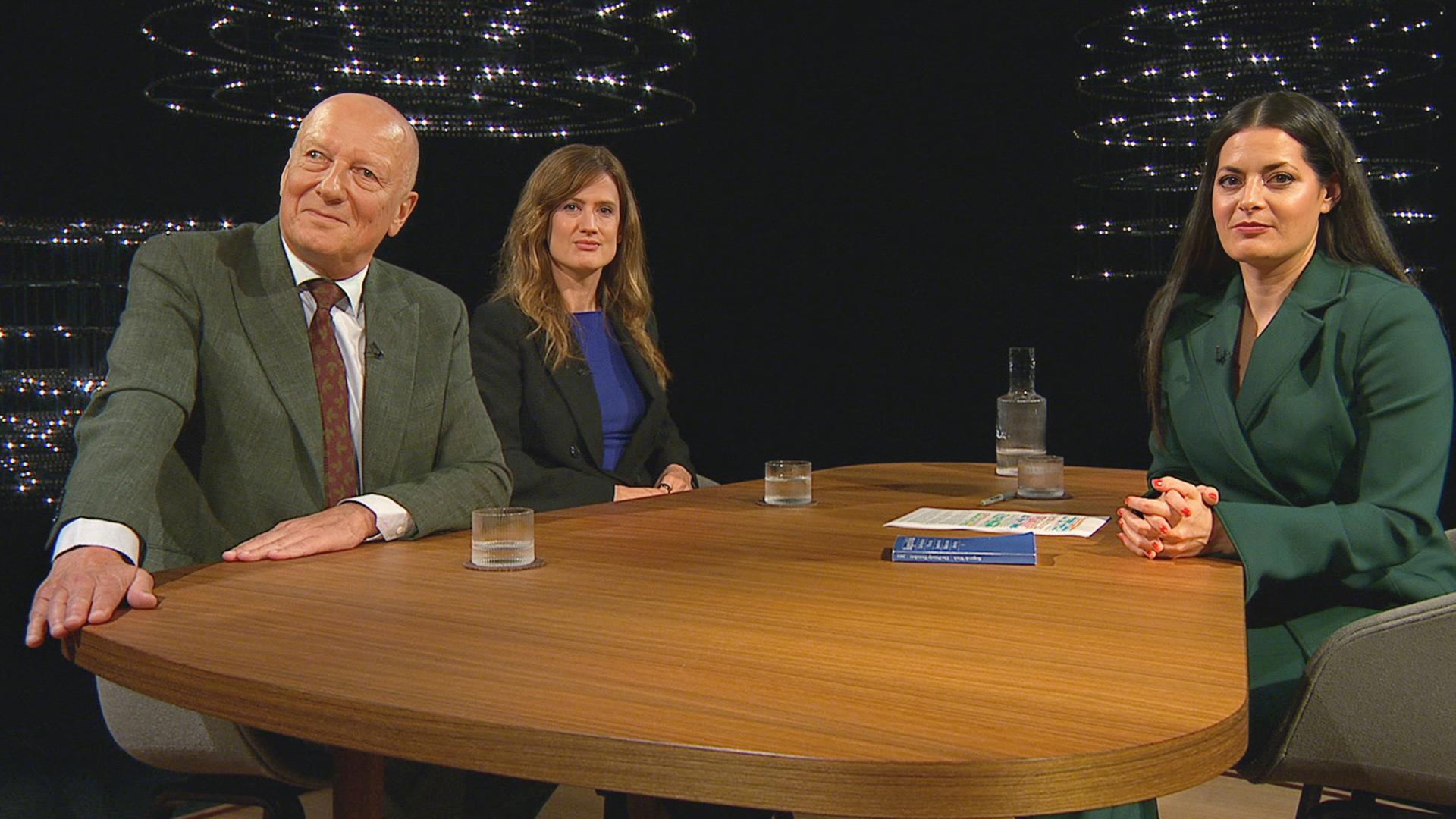 ADrei Personen sitzen an einem runden Holztisch in einem Fernsehstudio, zwei Frauen und ein Mann, vor dunklem Hintergrund und moderner Lichtinstallation.