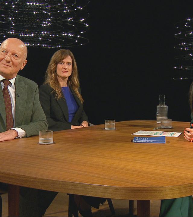 ADrei Personen sitzen an einem runden Holztisch in einem Fernsehstudio, zwei Frauen und ein Mann, vor dunklem Hintergrund und moderner Lichtinstallation.
