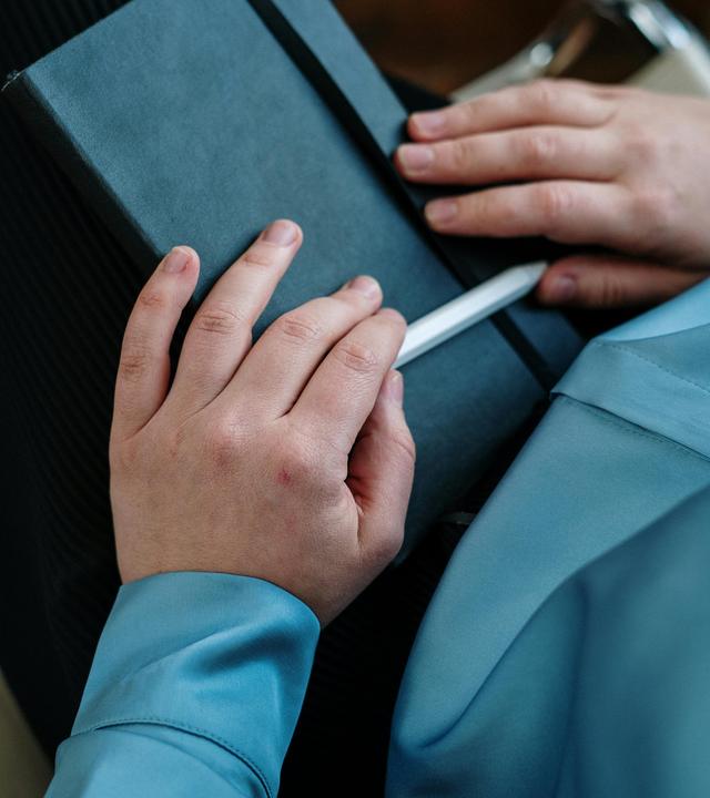Hände mit einem Stift in der Hand liegen auf einem Notizbuch im Schoß eines Menschen