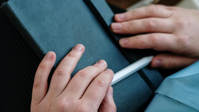 Hände mit einem Stift in der Hand liegen auf einem Notizbuch im Schoß eines Menschen