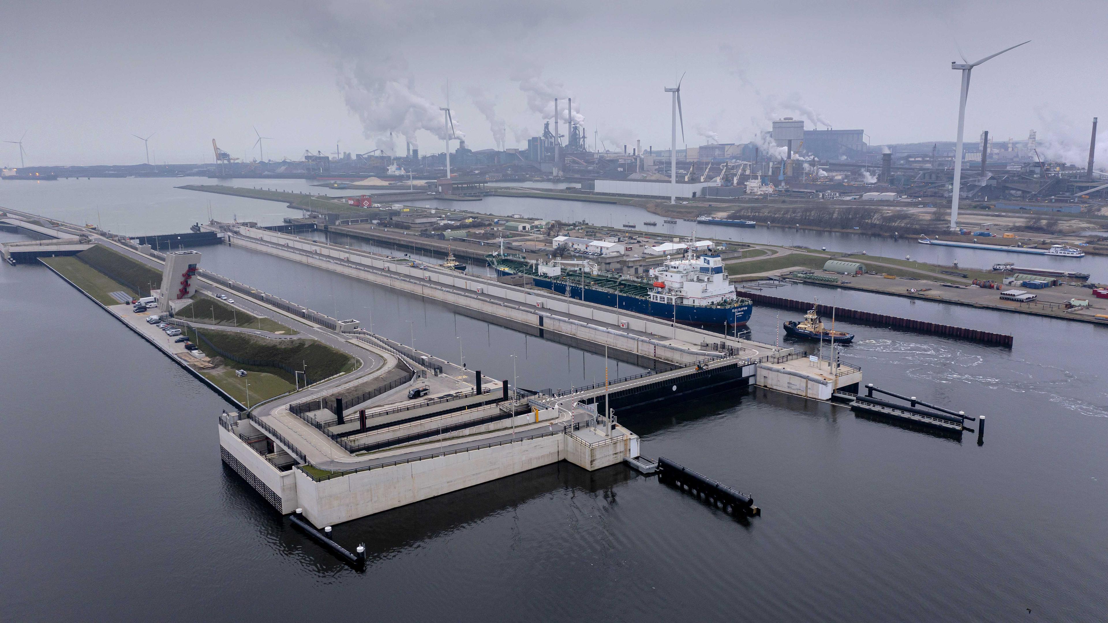 Eine riesige Schleuse im Wasser, darum herum ein Hafen und Industrie im Hintergrund.