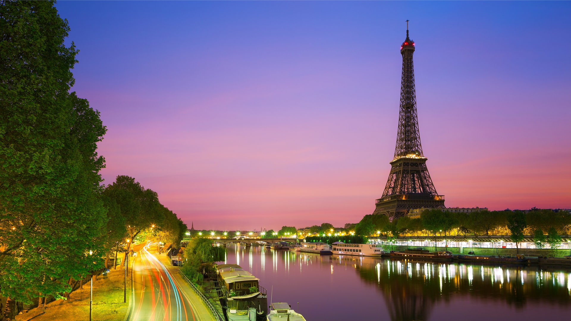 "Megacitys - Wenn es Nacht wird in Paris": Ein Fahrradfahrer fährt bei Nacht unter einer Brücke auf einem Radweg. Dahinter liegen die Lichter der Stadt Paris mit dem Eiffelturm.