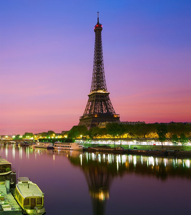 "Megacitys - Wenn es Nacht wird in Paris": Ein Fahrradfahrer fährt bei Nacht unter einer Brücke auf einem Radweg. Dahinter liegen die Lichter der Stadt Paris mit dem Eiffelturm.