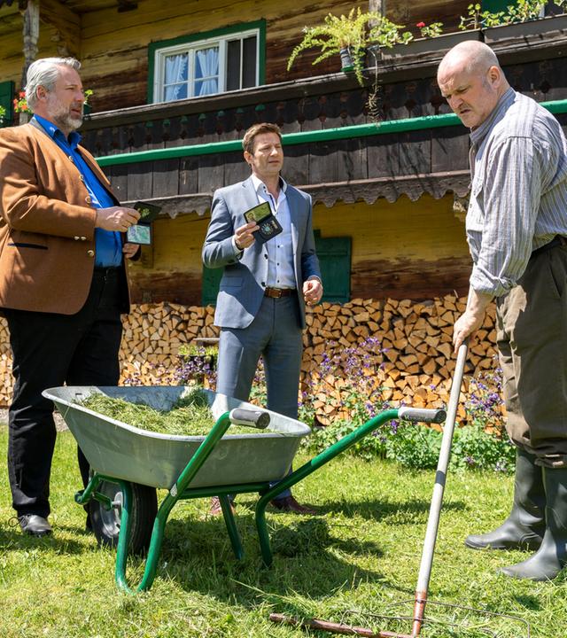 "Die Rosenheim-Cops - Mehr Schein als Sein": Die Kommissare Stadler (Dieter Fischer) und Hansen (Igor Jeftić) stehen auf einer Wiese vor einer Schubkarre, die mit Gras gefüllt ist, und zeigen Lukas Hirsch (Rainer Haustein), der Gras zusammenrecht, ihre Dienstmarke. Im Hintergrund ist ein bayerisches Holzhaus zu sehen, vor dessen Fenstern Holzscheite gestapelt sind.
