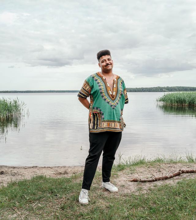 Dennis, ein junger Mann mit dunklem Teint, steht am Ufer eines ruhigen Sees, der Himmel ist größtenteils bewölkt. Er steht auf einer kleinen Grasfläche, die Hände sind locker in die Hosentaschen gesteckt, die Körperhaltung wirkt entspannt und freundlich.