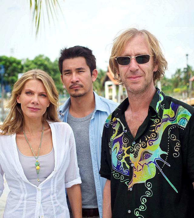 Eine blonde Frau steht mit zwei Männern an einem Boots-Steg. Einer der Männer hat asiatische Gesichtszüge, der andere trägt eine Sonnenbrille und wirkt europäisch.