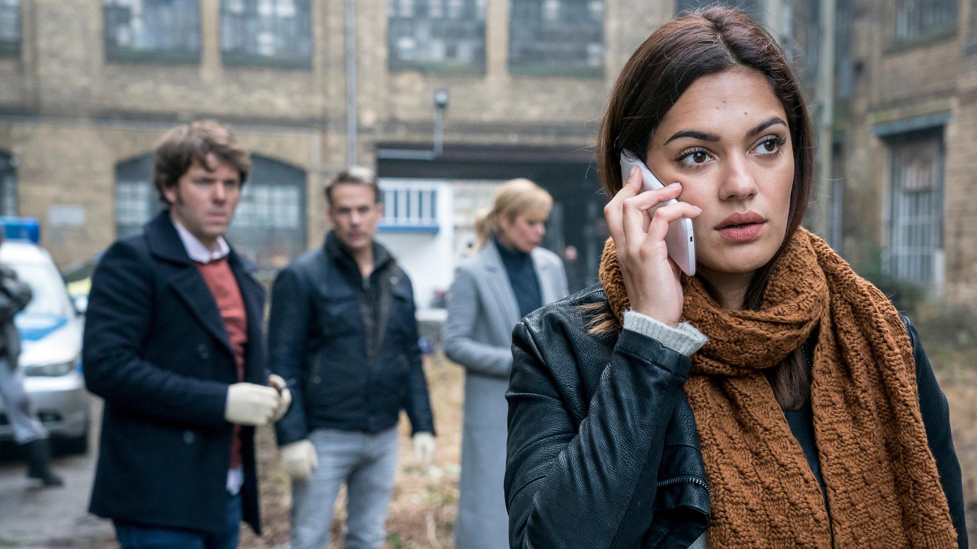 Olivia Fareedi steht im Hinterhof eines alten Gebäudes. Hinter ihr stehen ihre Kollegen und beobachten sie