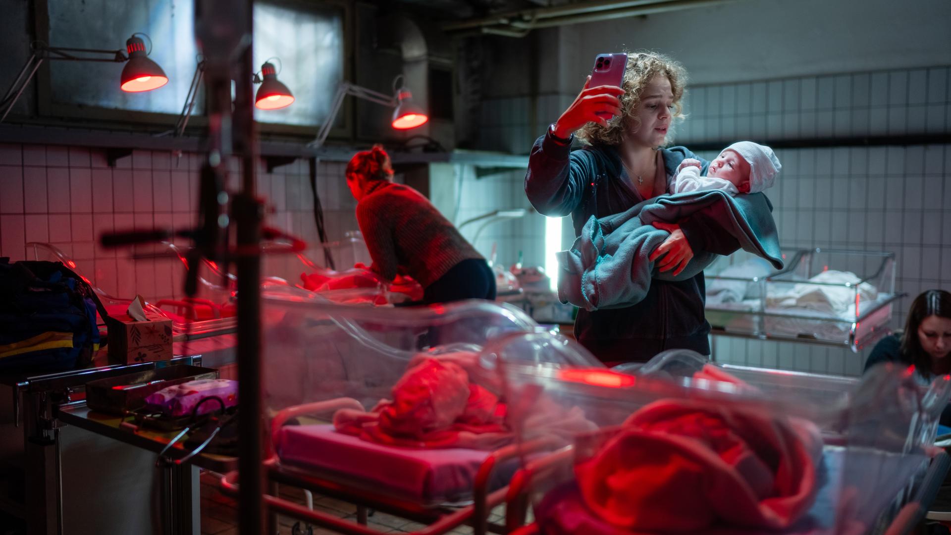  Oksana Smirnova (Alina Danko) steht in einer Überwachungsstation von Neugeborenen und hält ihr Baby auf dem linken Arm. In ihrer rechten Hand hält sie ihr Handy nach oben, um zu telefonieren. Um sie herum sind Wärmelampen, die auf die Neugeborene gerichtet sind.
