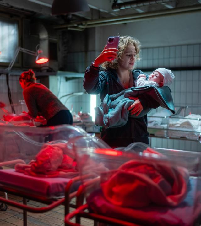  Oksana Smirnova (Alina Danko) steht in einer Überwachungsstation von Neugeborenen und hält ihr Baby auf dem linken Arm. In ihrer rechten Hand hält sie ihr Handy nach oben, um zu telefonieren. Um sie herum sind Wärmelampen, die auf die Neugeborene gerichtet sind.