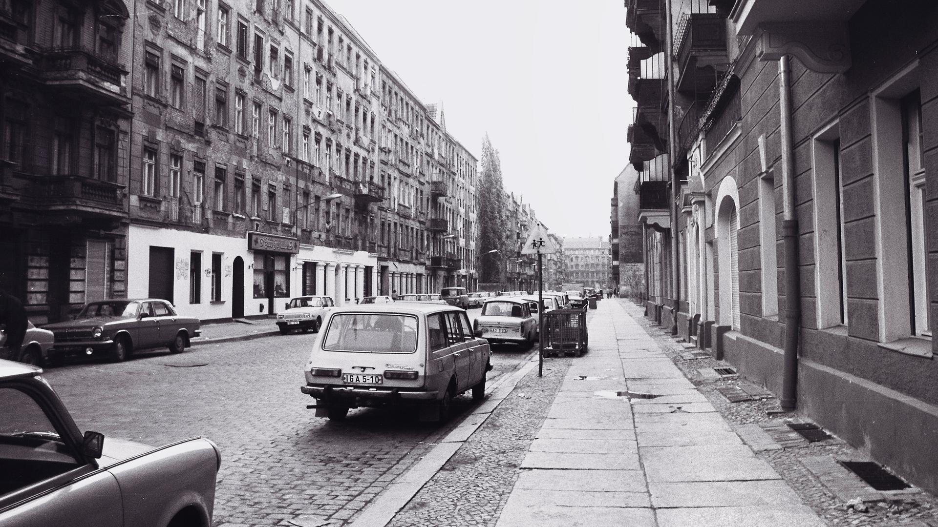 Schwarzweißfotografie einer städtischen Straßenszene in Ost-Berlin: Ende der 1980er Jahre. Links und rechts der Straße stehen mehrstöckige Altbauten, deren Fassaden teilweise verwittert wirken, mit bröckelndem Putz und Spuren von Abnutzung versehen sind. Die Straße ist mit Kopfsteinpflaster belegt, am rechten Rand verläuft ein breiter Gehweg mit großen Betonplatten auf denen sich Pfützen gebildet haben. Mehrere Automobile der Fabrikanten Trabant und Wartburg parken beidseitig entlang der Straße.