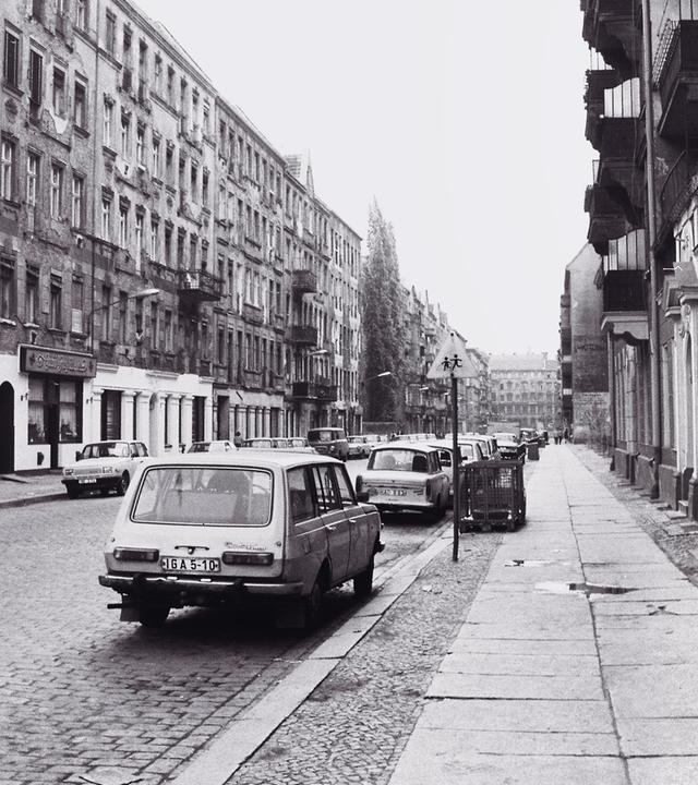 Schwarzweißfotografie einer städtischen Straßenszene in Ost-Berlin: Ende der 1980er Jahre. Links und rechts der Straße stehen mehrstöckige Altbauten, deren Fassaden teilweise verwittert wirken, mit bröckelndem Putz und Spuren von Abnutzung versehen sind. Die Straße ist mit Kopfsteinpflaster belegt, am rechten Rand verläuft ein breiter Gehweg mit großen Betonplatten auf denen sich Pfützen gebildet haben. Mehrere Automobile der Fabrikanten Trabant und Wartburg parken beidseitig entlang der Straße.
