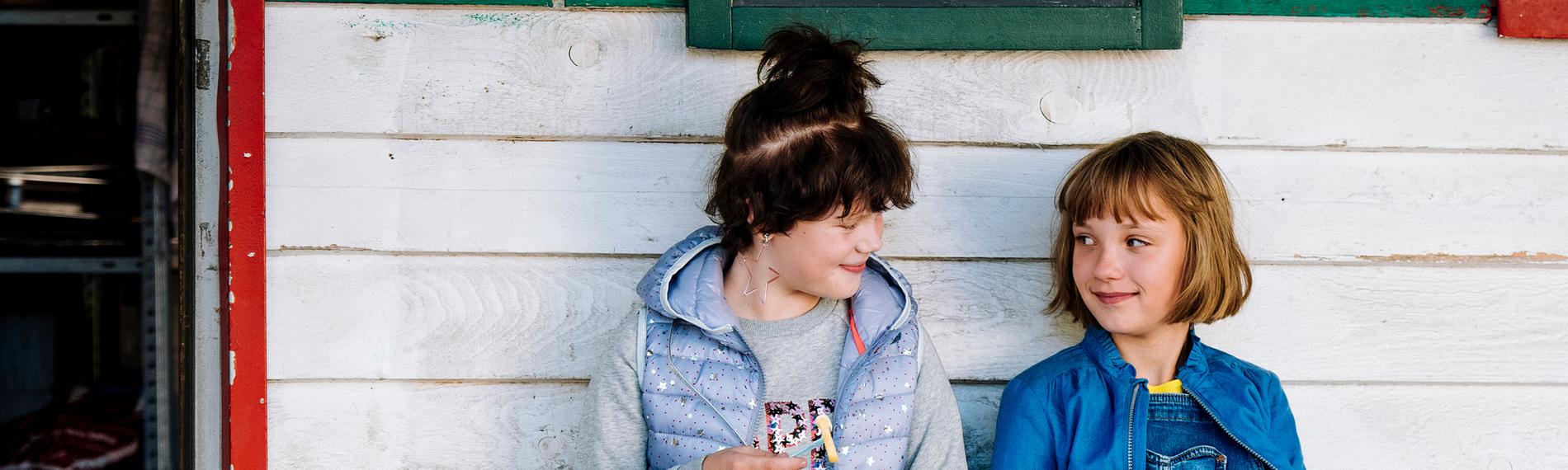 Cheyenne (Yola Streese) und Lotta (Meggy Hussong) sitzen auf einer Bank vor einer weiß-grün gestrichenen Hauswand neben der Eingangstür. Jede hält eine Portion Pommes in den Händen. Sie sehen sich lächelnd und zufrieden an.
