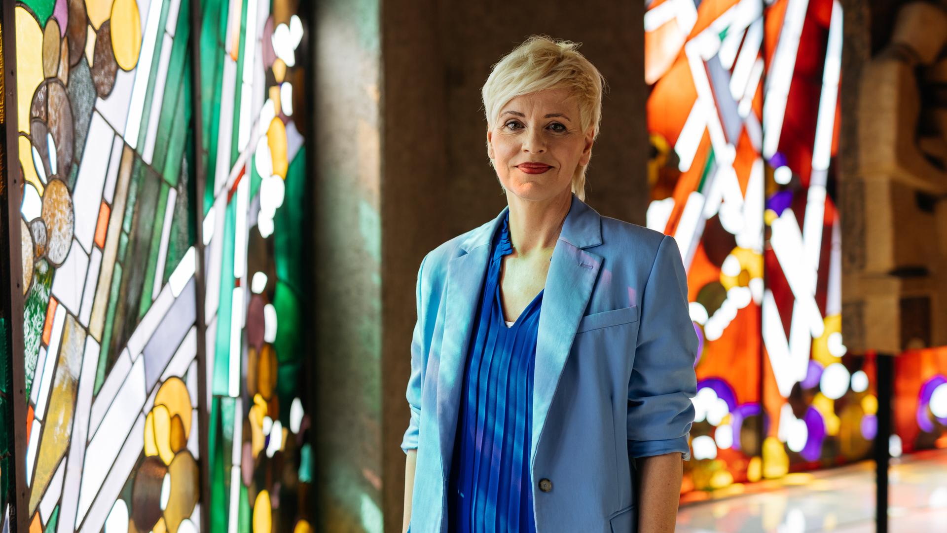 Christina steht in der Kirche und schaut ein wenig verschmitzt in die Kamera. Im Hintergrund sind bunte Kirchenfenster zu sehen. 
