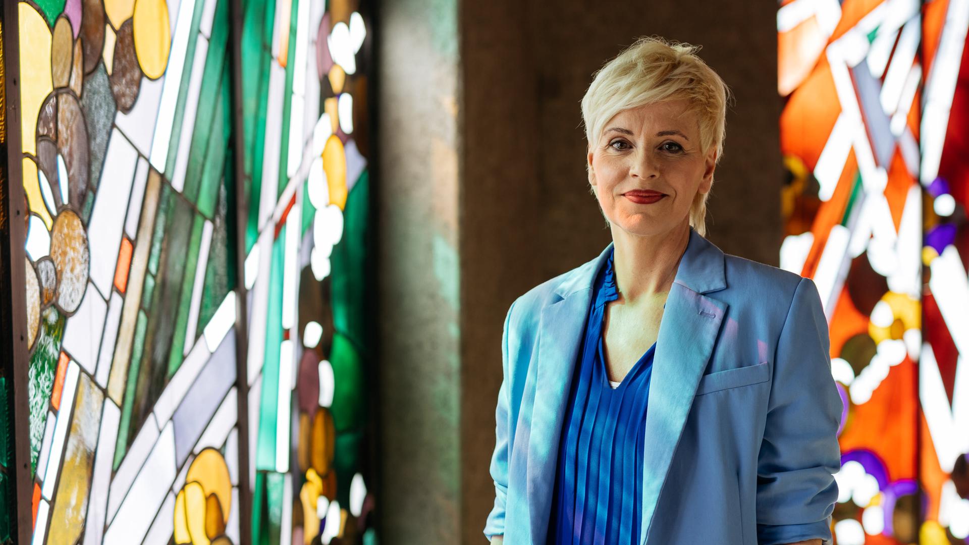 Christina steht in der Kirche und schaut ein wenig verschmitzt in die Kamera. Im Hintergrund sind bunte Kirchenfenster zu sehen. 