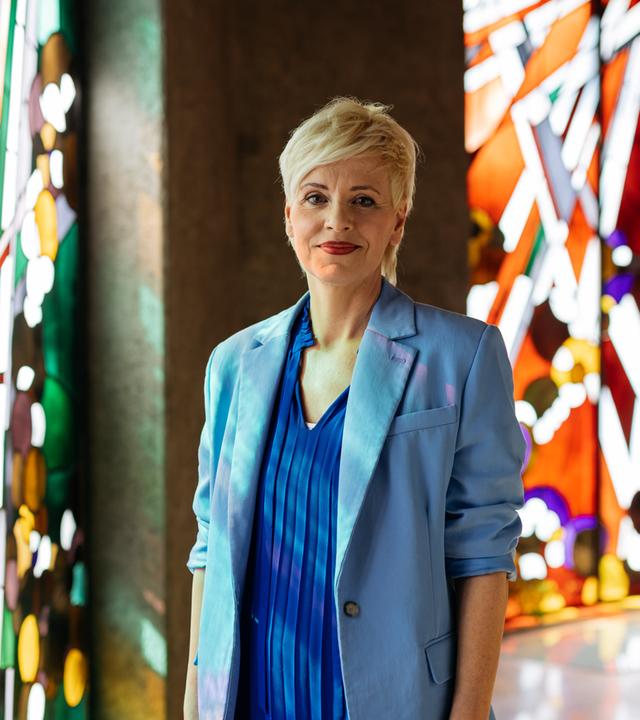 Christina steht in der Kirche und schaut ein wenig verschmitzt in die Kamera. Im Hintergrund sind bunte Kirchenfenster zu sehen. 