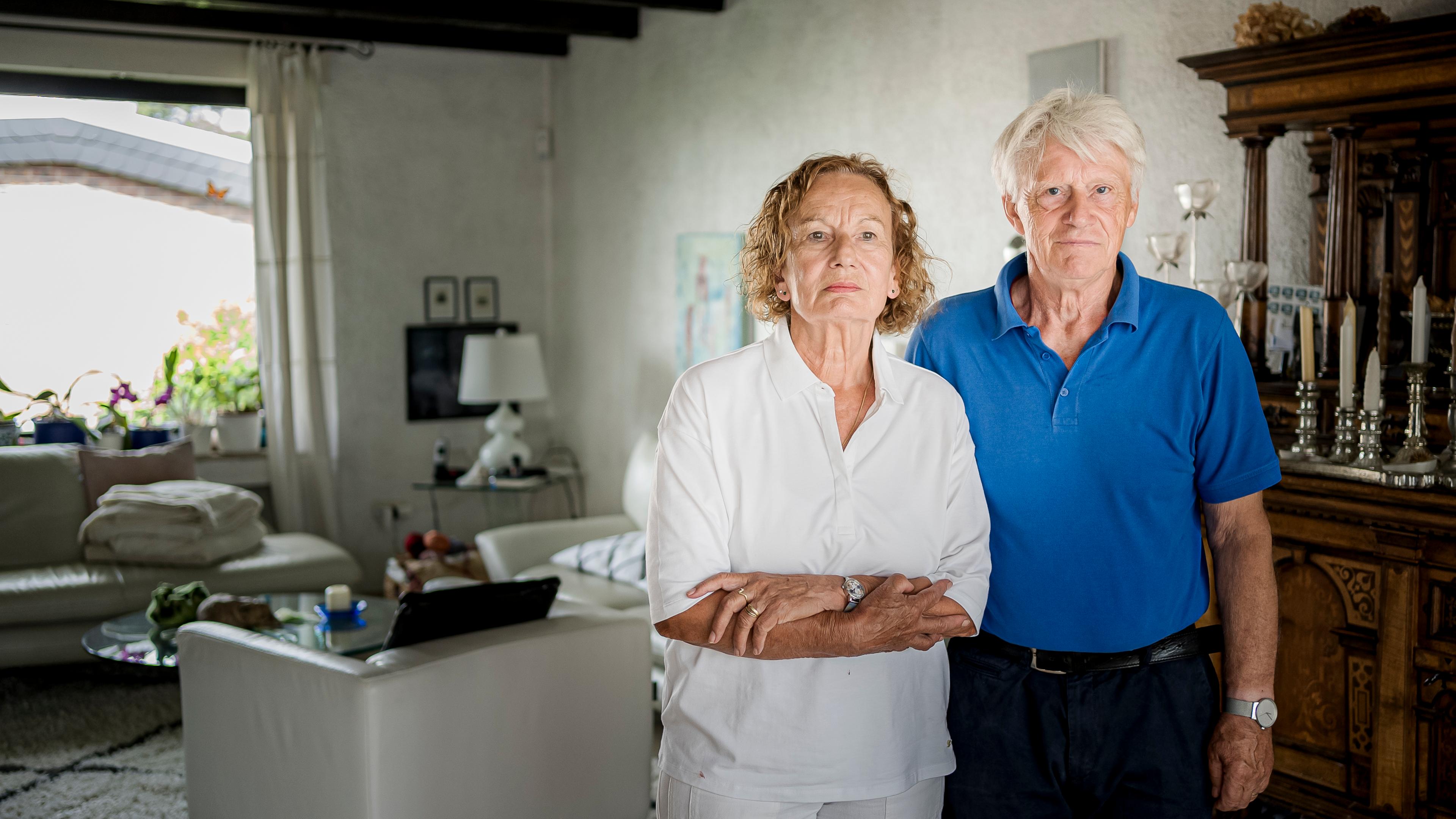 Sylvia und Armin Joos stehen im Wohnzimmer vor eine weißer Wand. Sylvia hat gelocktes kinnlanges Haar und trägt eine weiße Bluse, Armin hat graue Haare und trägt ein blaues Polo-Shirt.