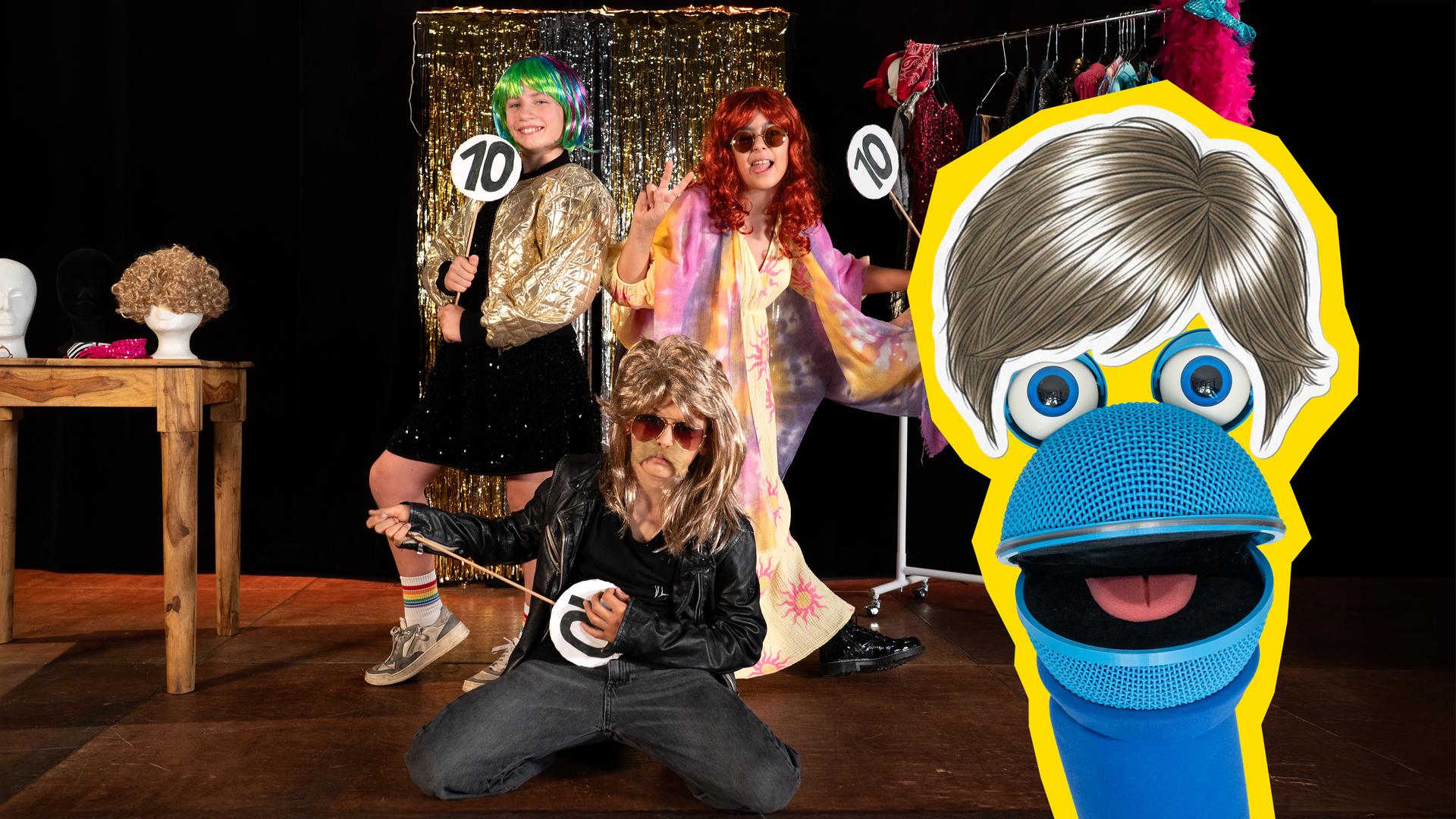 SingDing trägt eine Perücke, während drei verkleidete Kinder im Hintergrund Bewertungstafeln mit der Zahl 10 hochhalten. Im Hintergrund sieht man eine Kleiderstange mit bunten Kostümen, ein Lametta-Vorhang und einen Tisch, auf dem ein Perückenkopf steht.