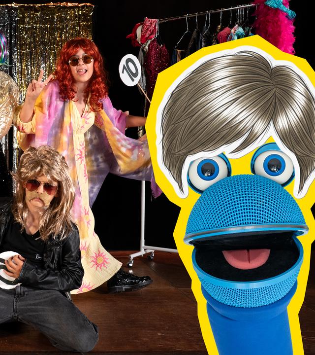 SingDing trägt eine Perücke, während drei verkleidete Kinder im Hintergrund Bewertungstafeln mit der Zahl 10 hochhalten. Im Hintergrund sieht man eine Kleiderstange mit bunten Kostümen, ein Lametta-Vorhang und einen Tisch, auf dem ein Perückenkopf steht.