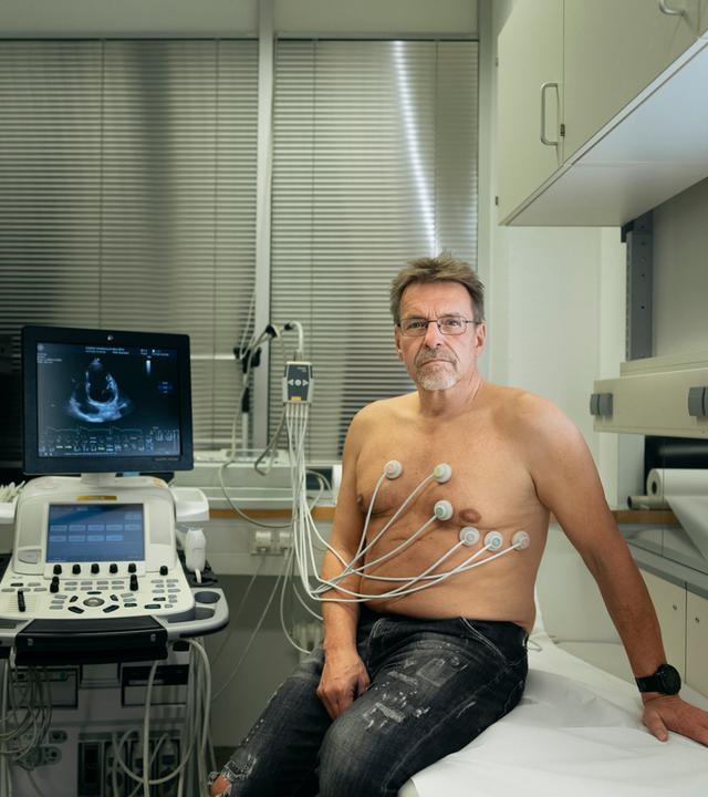 Harald sitzt auf einer Untersuchungsliege beim Arzt. Elektroden kleben auf seinem nackten Oberkörper. Auf dem Monitor neben ihm sieht man ein Ultraschallbild seines Herzens.