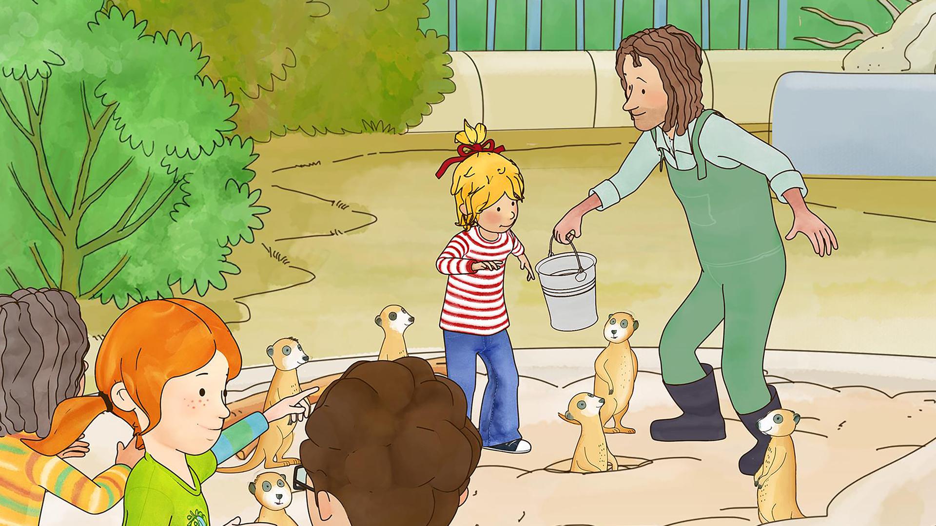Conni füttert die Erdmännchen im Zoo. Sie steht mit einem Tierpfleger im Gehege und greift nach dem Futter in einem Eimer. Im Vordergrund beobachten Kinder das Spektakel.