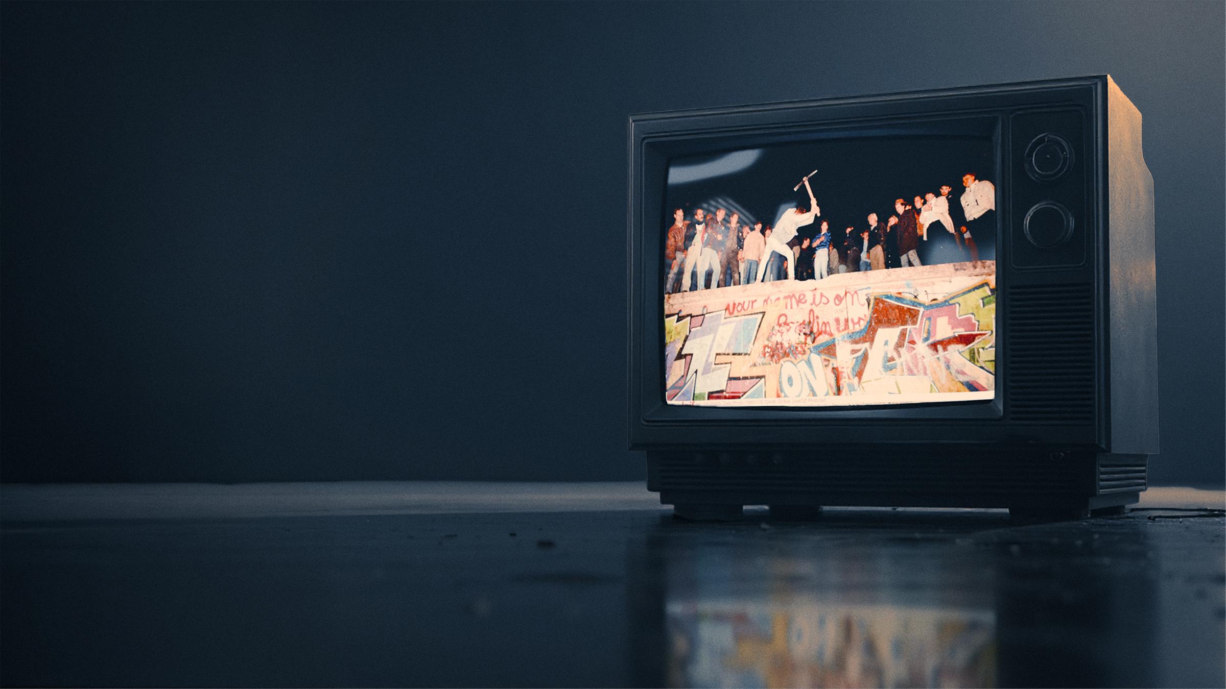 Ein alter Röhrenfernseher, der den Sturz der mauer zeigt. Auf ihr stehen mehrere Personen und feiern die Wiedervereinigung. 