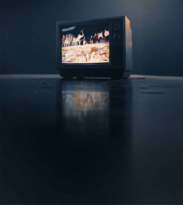 Ein alter Röhrenfernseher, der den Sturz der mauer zeigt. Auf ihr stehen mehrere Personen und feiern die Wiedervereinigung. 