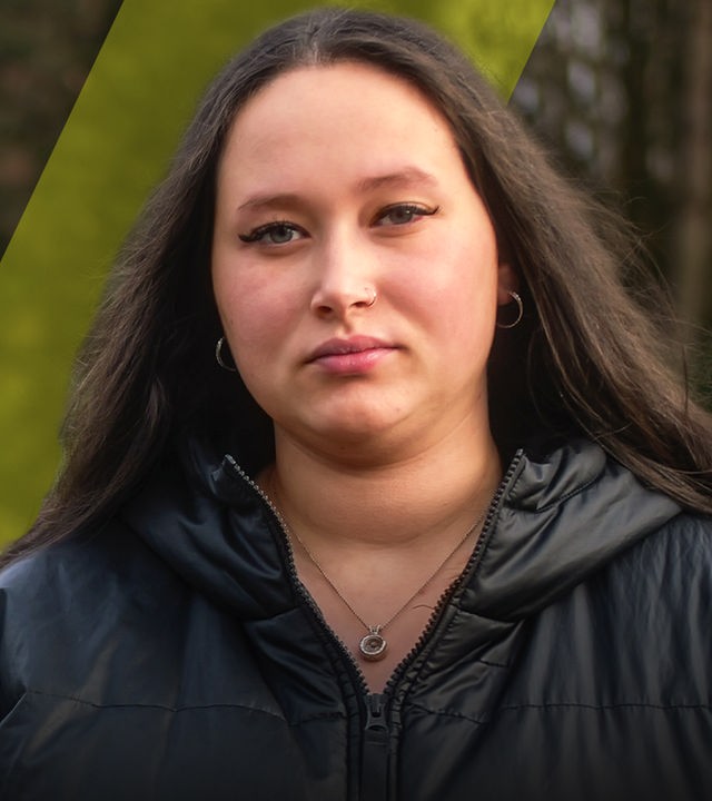 Eine junge Frau schaut traurig nach vorne. Im Hintergrund ist ein Friedhof zu sehen. 