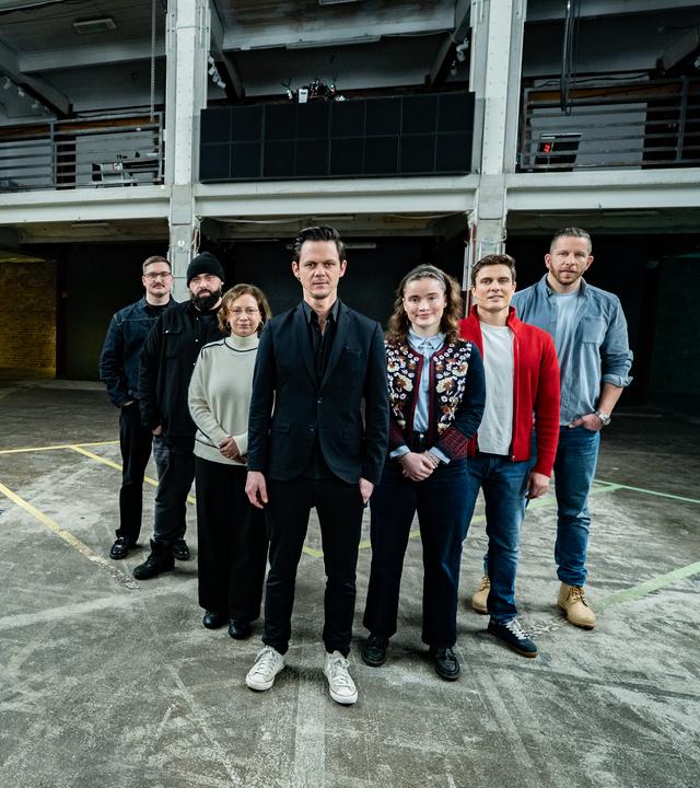 Es stehen sieben Menschen nebeneinander auf einem Boden, der ein schachbrettartiges Muster hat. Es sind zwei Frauen und fünf Männer. Alle blicken in die Richtung des Betrachters.