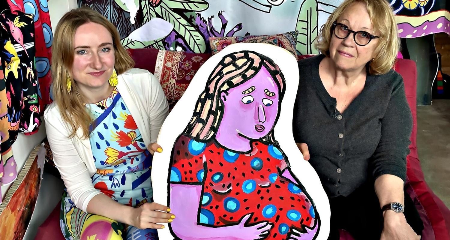 Zwei Frauen sitzen nebeneinander auf einem Sofa und halten gemeinsam ein farbenfrohes Gemälde einer lila Frau mit rotem Kleid; der Hintergrund ist mit bunten Mustern dekoriert.