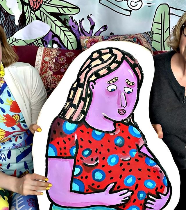 Zwei Frauen sitzen nebeneinander auf einem Sofa und halten gemeinsam ein farbenfrohes Gemälde einer lila Frau mit rotem Kleid; der Hintergrund ist mit bunten Mustern dekoriert.