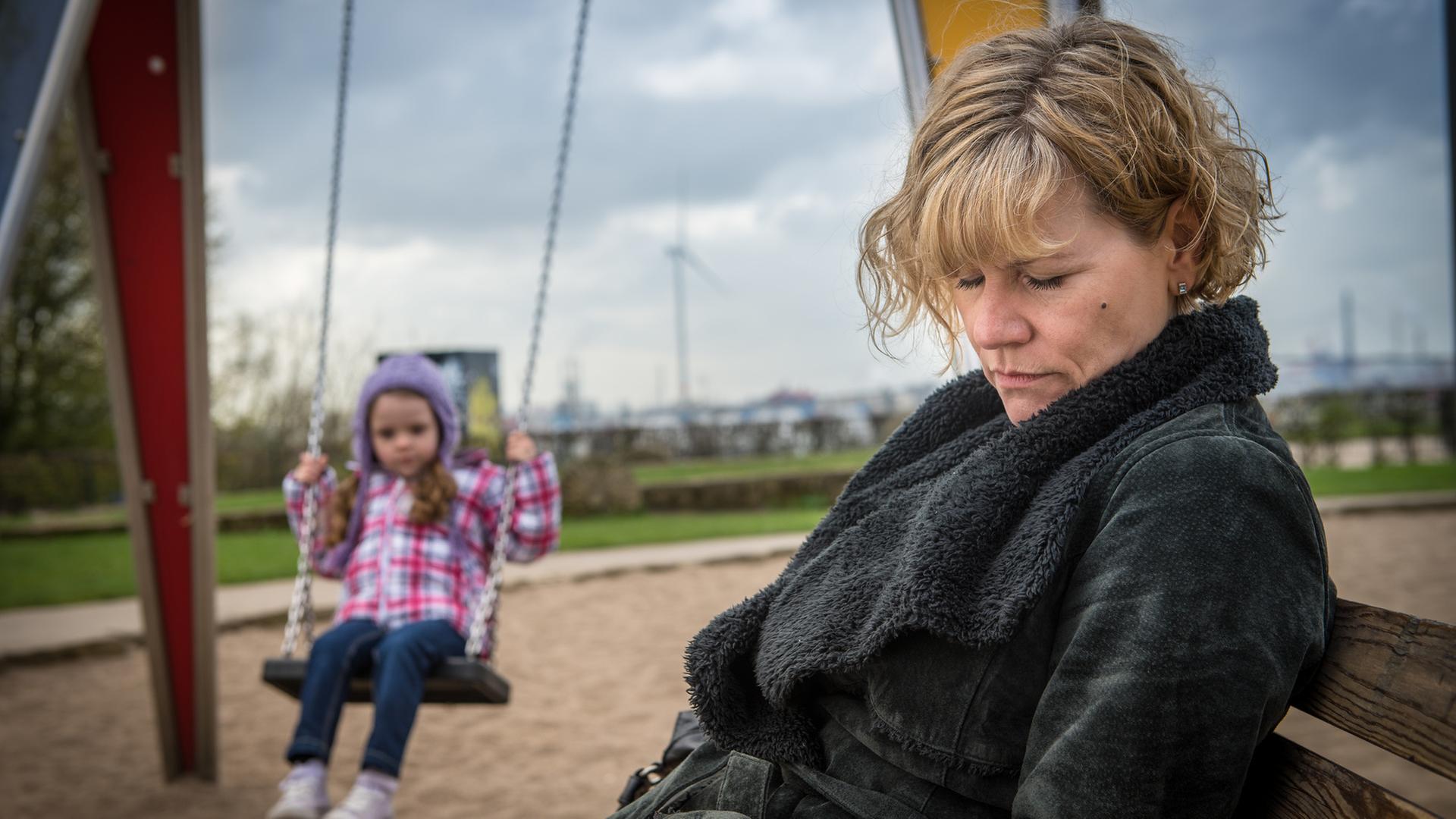 "Notruf Hafenkante - Melody": Im Vordergrund sitzt Karin Berger (Anne Kasprik) schlafend auf einer Parkbank. Im Hintergrund schaukelt die fünfjährige Antonia (Neala Hoppe) auf einer Spielplatzschaukel.