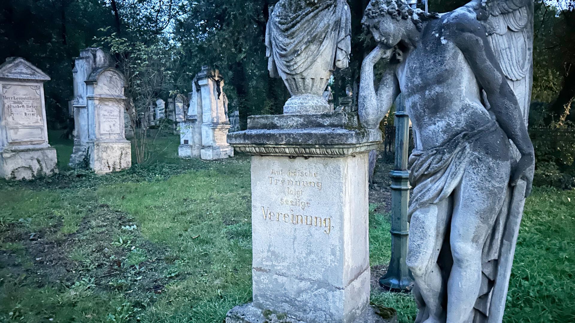 Das Bild zeigt eine Szene auf einem Friedhof, wahrscheinlich am Friedhof St. Marx. Im Vordergrund steht eine detaillierte Skulptur eines Engels, der nachdenklich mit einer Hand an seinem Kopf lehnt. Die Statue ist aus Stein und wirkt verziert und alt. Neben dem Engel befindet sich ein monumentaler Grabstein mit der Inschrift „Auferstehung“ in goldener Schrift.   Im Hintergrund sind mehrere Grabsteine zu sehen, die unterschiedlich geformt und angeordnet sind, und die Grabinschriften sind teilweise nicht klar lesbar. Die Umgebung ist grün, mit Gras und einigen Bäumen, die eine ruhige und friedliche Atmosphäre schaffen. Es scheint Dämmerung zu sein, was dem Bild eine melancholische Stimmung verleiht.   Die Gesamtszene vermittelt ein Gefühl von Trauer, aber auch von Trost durch die künstlerische Darstellung des Engels und den historischen Kontext des Ortes.
