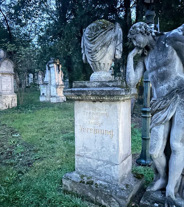 Das Bild zeigt eine Szene auf einem Friedhof, wahrscheinlich am Friedhof St. Marx. Im Vordergrund steht eine detaillierte Skulptur eines Engels, der nachdenklich mit einer Hand an seinem Kopf lehnt. Die Statue ist aus Stein und wirkt verziert und alt. Neben dem Engel befindet sich ein monumentaler Grabstein mit der Inschrift „Auferstehung“ in goldener Schrift.   Im Hintergrund sind mehrere Grabsteine zu sehen, die unterschiedlich geformt und angeordnet sind, und die Grabinschriften sind teilweise nicht klar lesbar. Die Umgebung ist grün, mit Gras und einigen Bäumen, die eine ruhige und friedliche Atmosphäre schaffen. Es scheint Dämmerung zu sein, was dem Bild eine melancholische Stimmung verleiht.   Die Gesamtszene vermittelt ein Gefühl von Trauer, aber auch von Trost durch die künstlerische Darstellung des Engels und den historischen Kontext des Ortes.